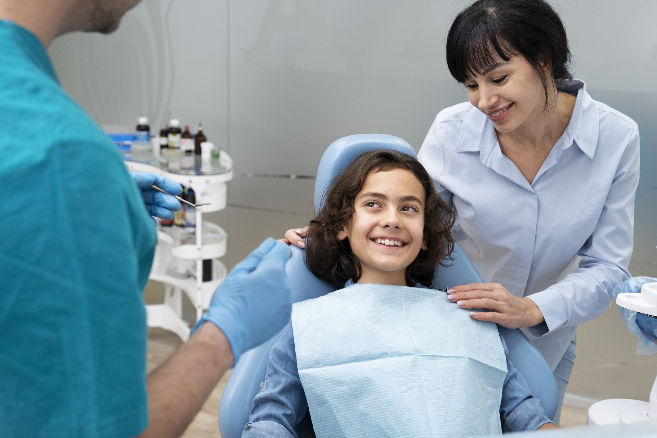 A smiling child sits in a dental chair wearing a bib while a dentist in gloves prepares an instrument, and a woman stands beside the child with a reassuring hand on their shoulder in a dental clinic.