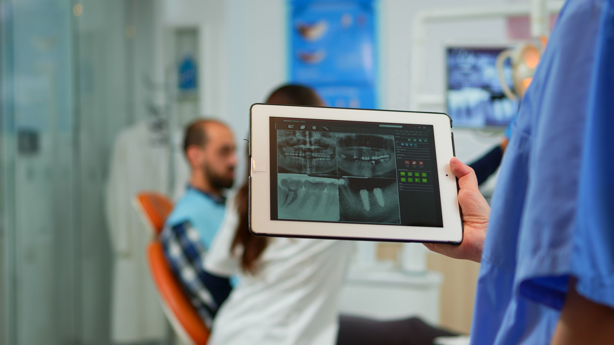 Dentist reviewing digital dental X-rays on a tablet while a patient sits in a dental chair during an orthodontic consultation.Dentist reviewing digital dental X-rays on a tablet while a patient sits in a dental chair during an orthodontic consultation.
