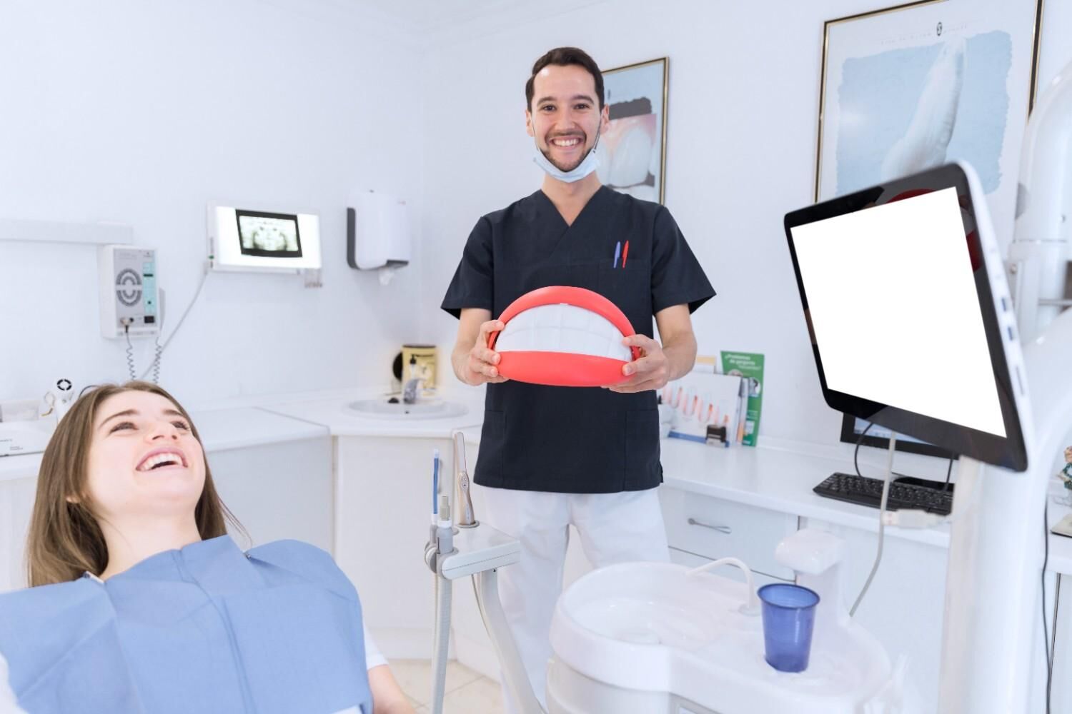 A smiling dentist holds a large tooth model while a patient reclines in a dental chair in a bright clinic.